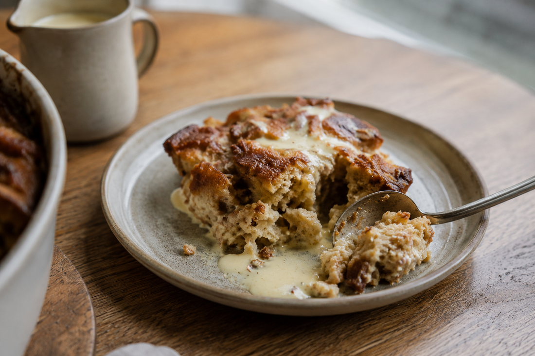 A serving of masala chai bread and butter pudding with cream on a wooden table beside a rainy window