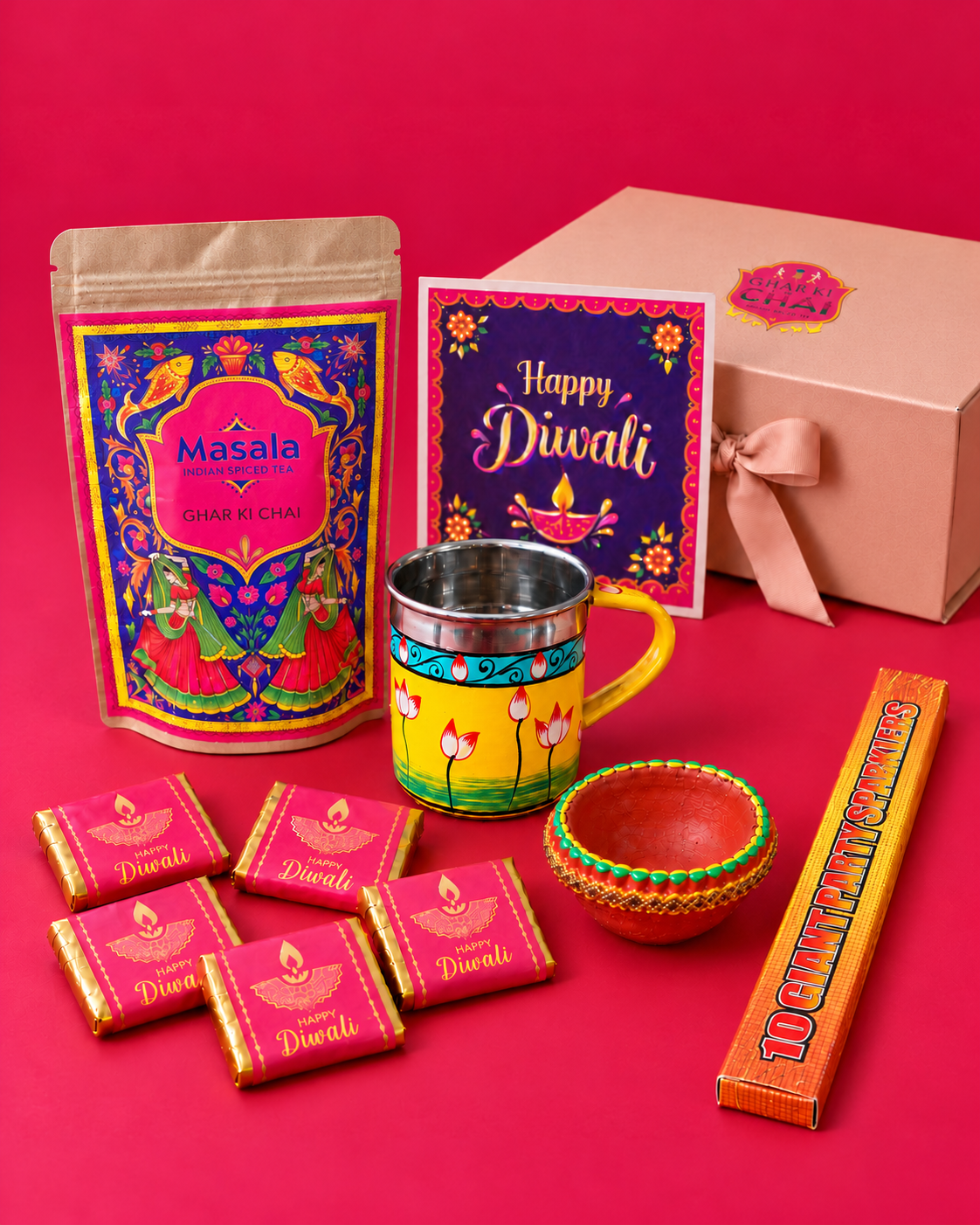 Diwali gift box contents arranged on a pink-red backdrop beside the blush pink gift box